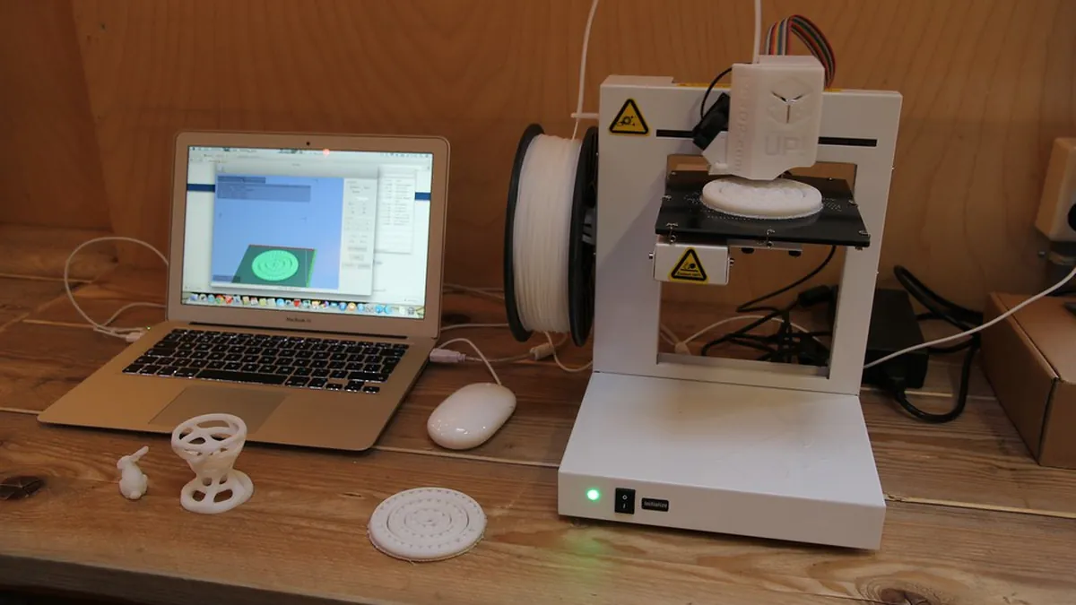 A white 3D printer with a filament spool mounted, next to a laptop and several printed parts.