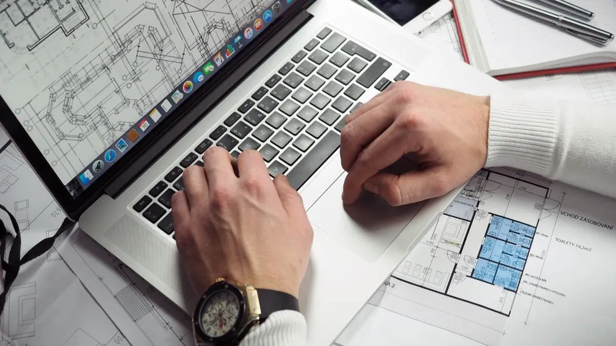 Hands typing on a laptop on a desk covered in technical drawings.
